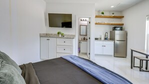A bedroom with a bed, a TV mounted on the wall, a kitchen area with a refrigerator, microwave, and wooden shelves. - Easy South Padre Beach Retreat w/ Pool Access (South Padre Island)