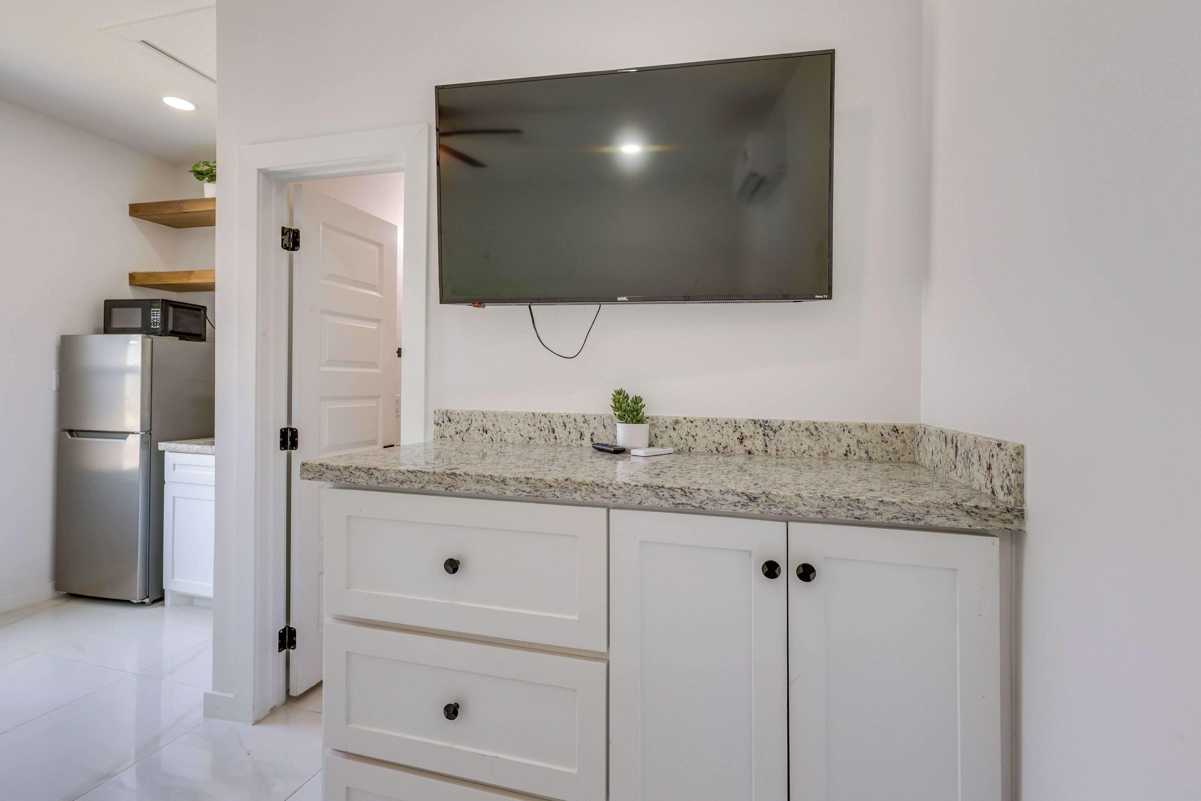A modern kitchen with white cabinets, a granite countertop, a flat-screen TV, and a stainless steel refrigerator.