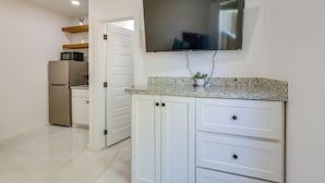 Una cocina con armarios blancos, encimera de granito, microondas, refrigerador y televisor de pantalla plana montado en la pared.