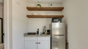 Eine kleine Küche mit weißen Schränken, einem Edelstahlkühlschrank und einem Holzregal darüber.