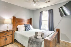 Ein Schlafzimmer mit Holz-Kopfteil, einem Nachttisch mit Lampe, einem Bett mit Kissen, einem Fenster mit Vorhängen und einem am Wand montierten Flachbildfernseher.
