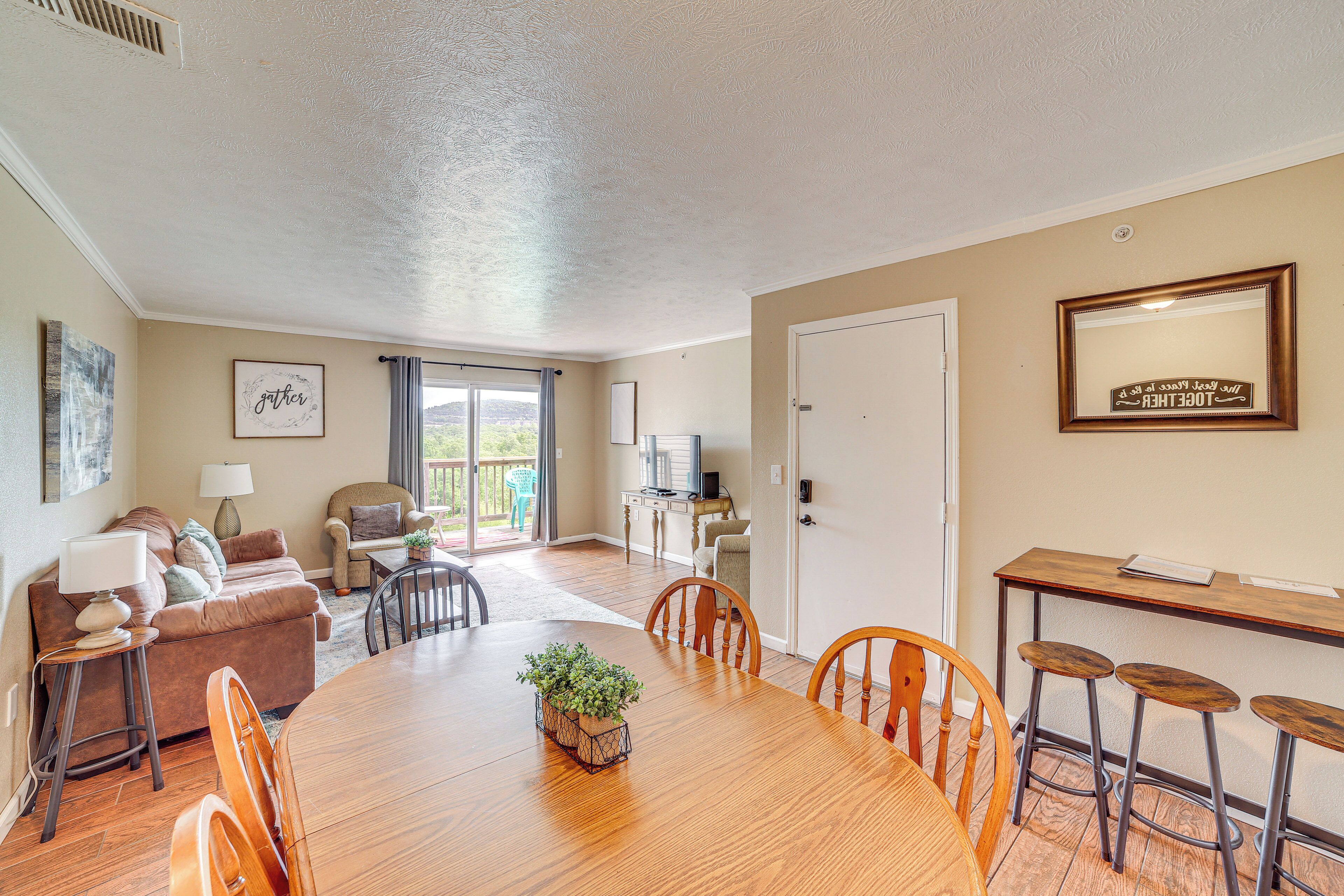 A spacious living area with a wooden dining table, comfortable seating, a television, and a balcony with a view.