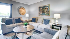 A living room with a beige sofa, a round coffee table, and a side table with a lamp. There is a painting on the wall and a window with curtains. - 3 Mi to Branson Strip: 2-unit Gem w/ Pool Access! (Branson)