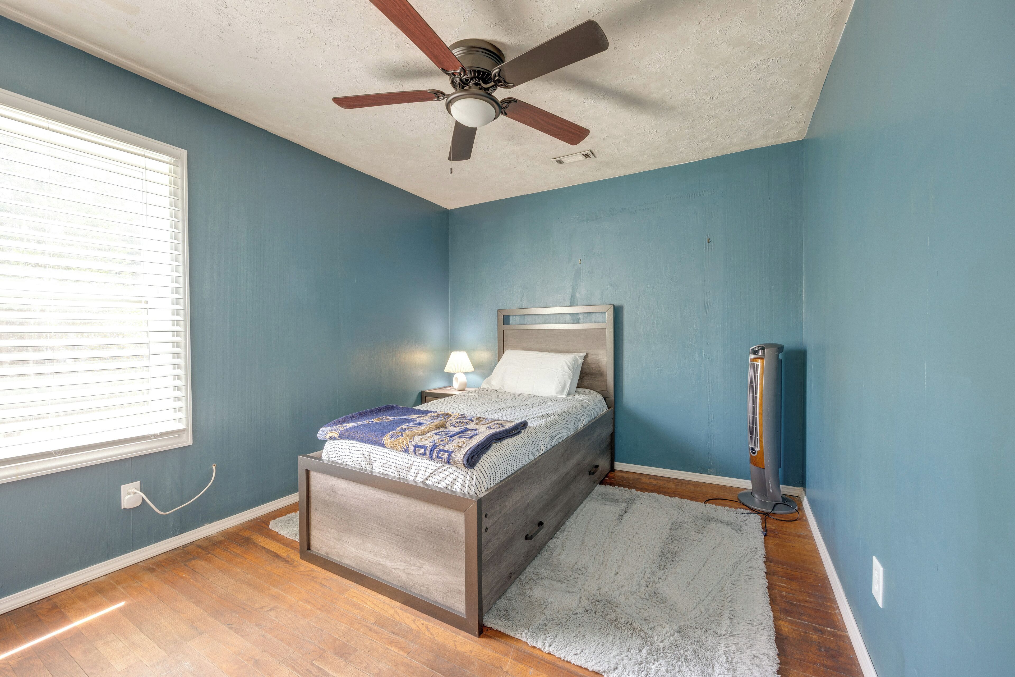 A bedroom with a bed, a ceiling fan, a standing heater, and a window with blinds.