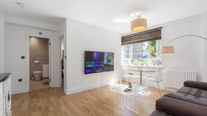 Uma sala de estar moderna com uma TV de tela plana, uma mesa de centro de vidro, um sofá marrom e uma área de jantar com mesa e cadeiras.