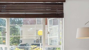 Uma área de refeições moderna com uma mesa de vidro redonda, cadeiras transparentes, base de metal, uma janela com persianas e um candeeiro suspenso.