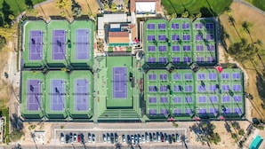 Vista aérea de un complejo deportivo con varias canchas de tenis, áreas de estacionamiento y un edificio central.