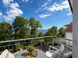 A balcony with a view of the sea, surrounded by trees and equipped with outdoor furniture.
