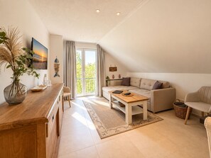 A modern living room with a sofa, a coffee table, a dining area with a wooden table and chairs, and a large window with curtains. - Oceanview Residences Lauterbach Rügen (Putbus)