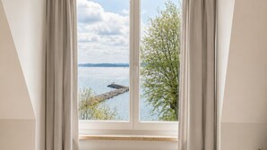 A room with a large window, white curtains, a radiator, and a view of a body of water and trees.