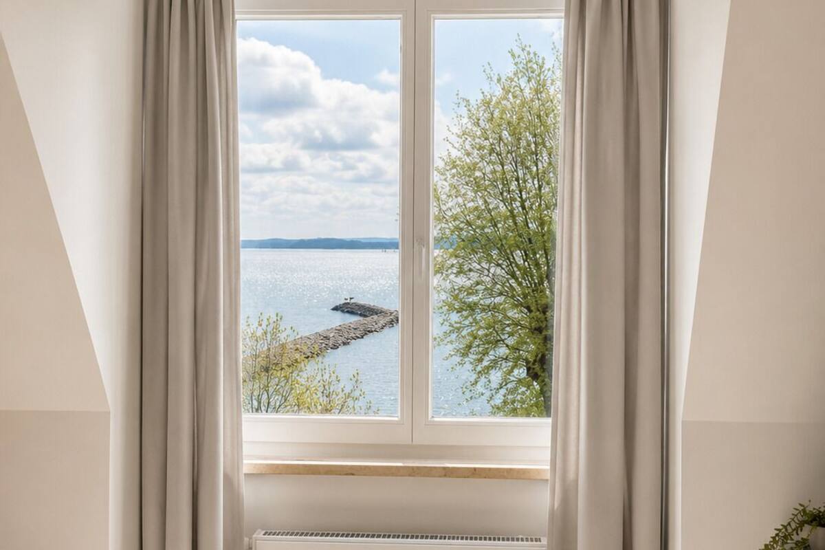 Una habitación con un ventanal grande, cortinas blancas, un radiador y vistas a un cuerpo de agua y árboles.