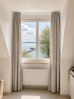 Una habitación con un ventanal grande, cortinas blancas, un radiador y vistas a un cuerpo de agua y árboles.