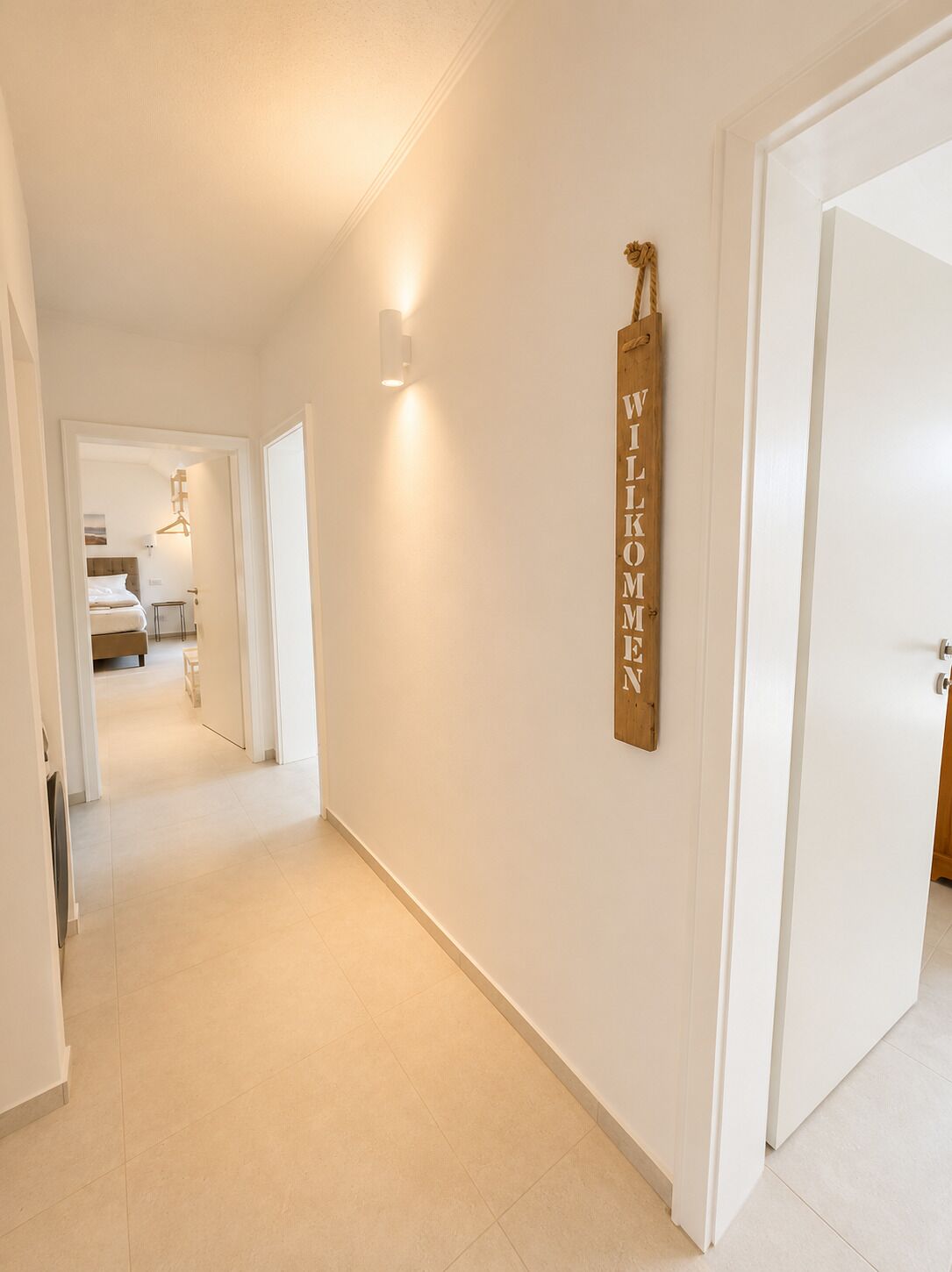 Un couloir avec une pancarte en bois sur le mur où est écrit « BIENVENUE », une porte sur la droite et un lit visible au fond.