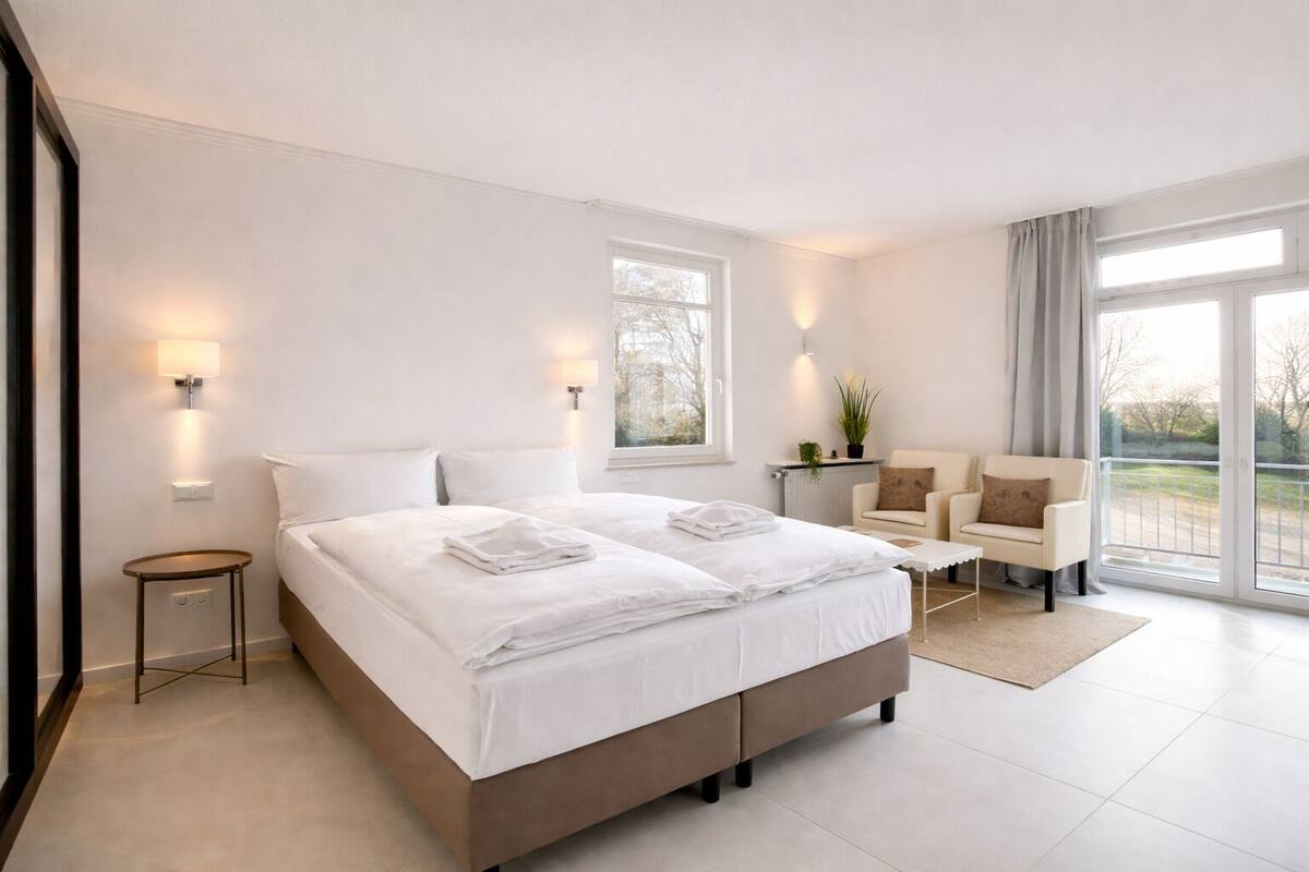 Una habitación de hotel moderna con una cama grande, un área de descanso con una silla, una mesita y vistas a través de un ventanal.