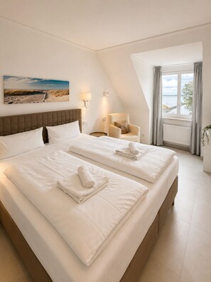 A hotel room with two beds, a view of the beach, a chair, and a window with curtains. - Oceanview Residences Lauterbach Rügen (Putbus)