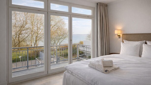 A hotel room with a large window, a bed with white linens, and a view of trees and water.