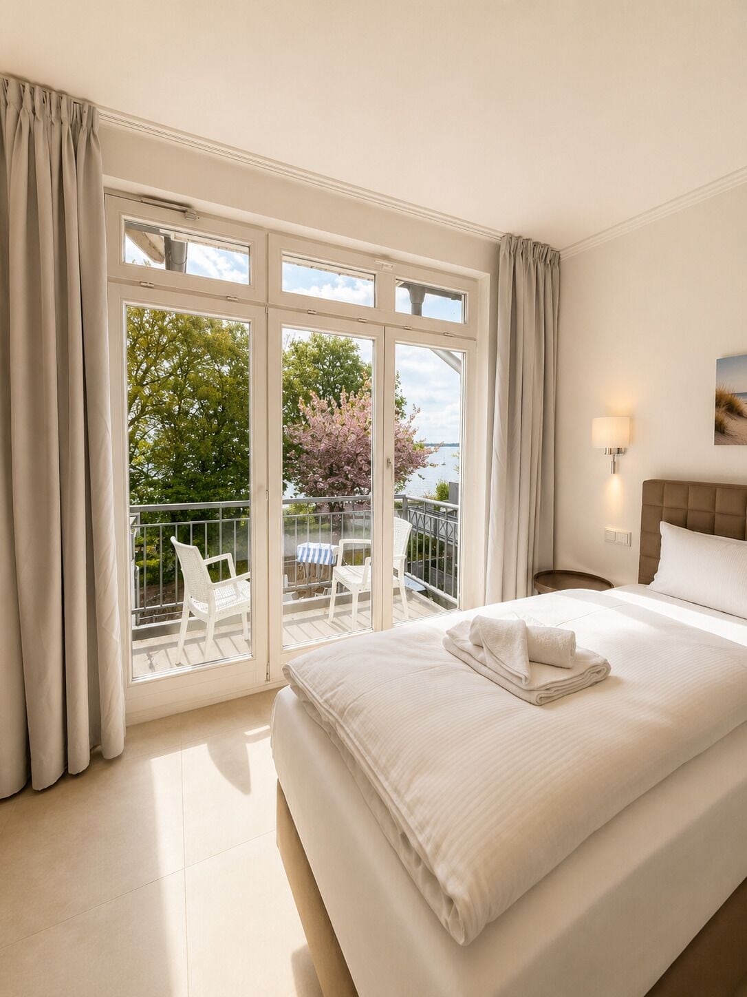 A hotel room with a large bed, a balcony with chairs, and a view of trees and water.