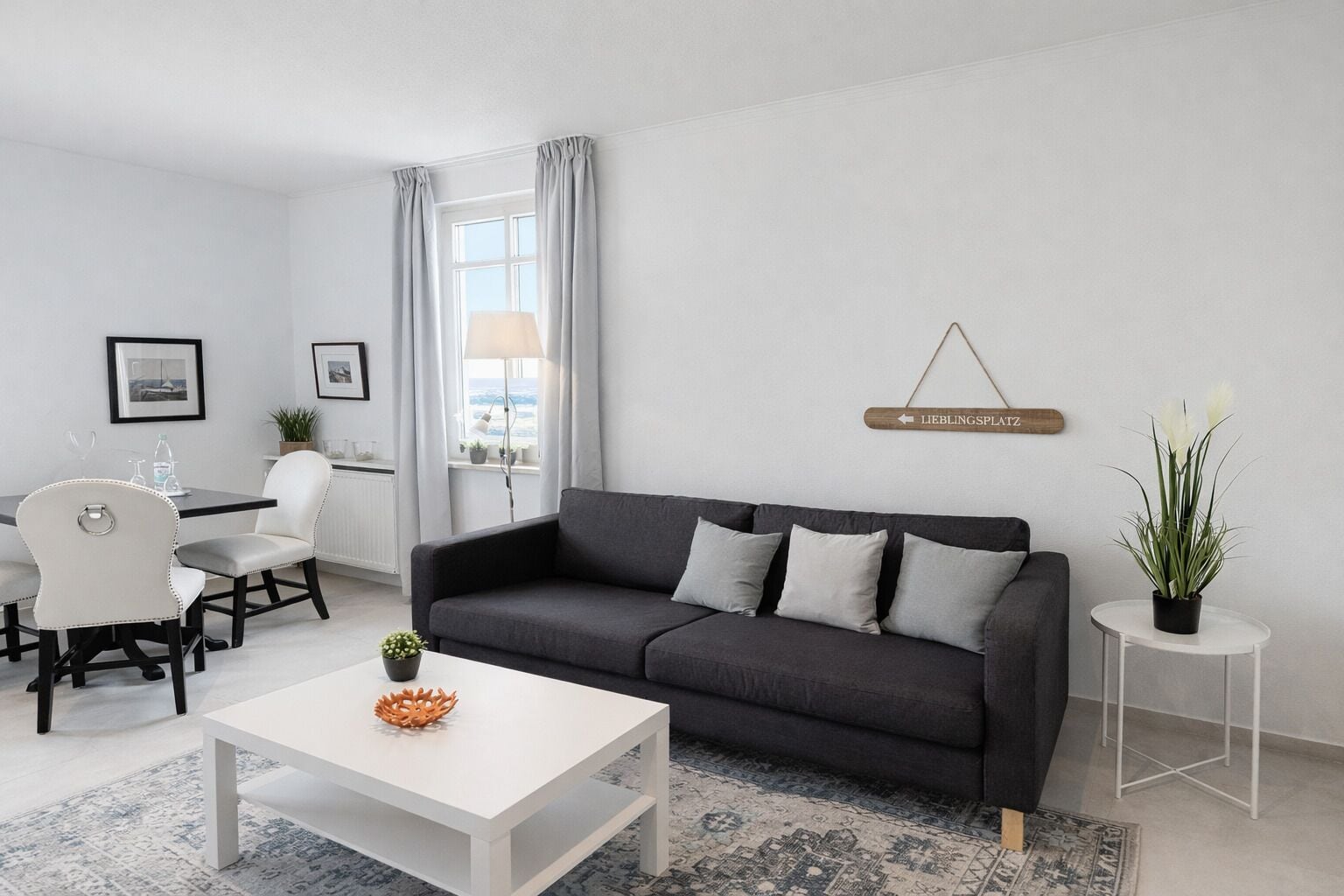A modern living room with a grey sofa, white coffee table, and a dining area with a white table and chairs.