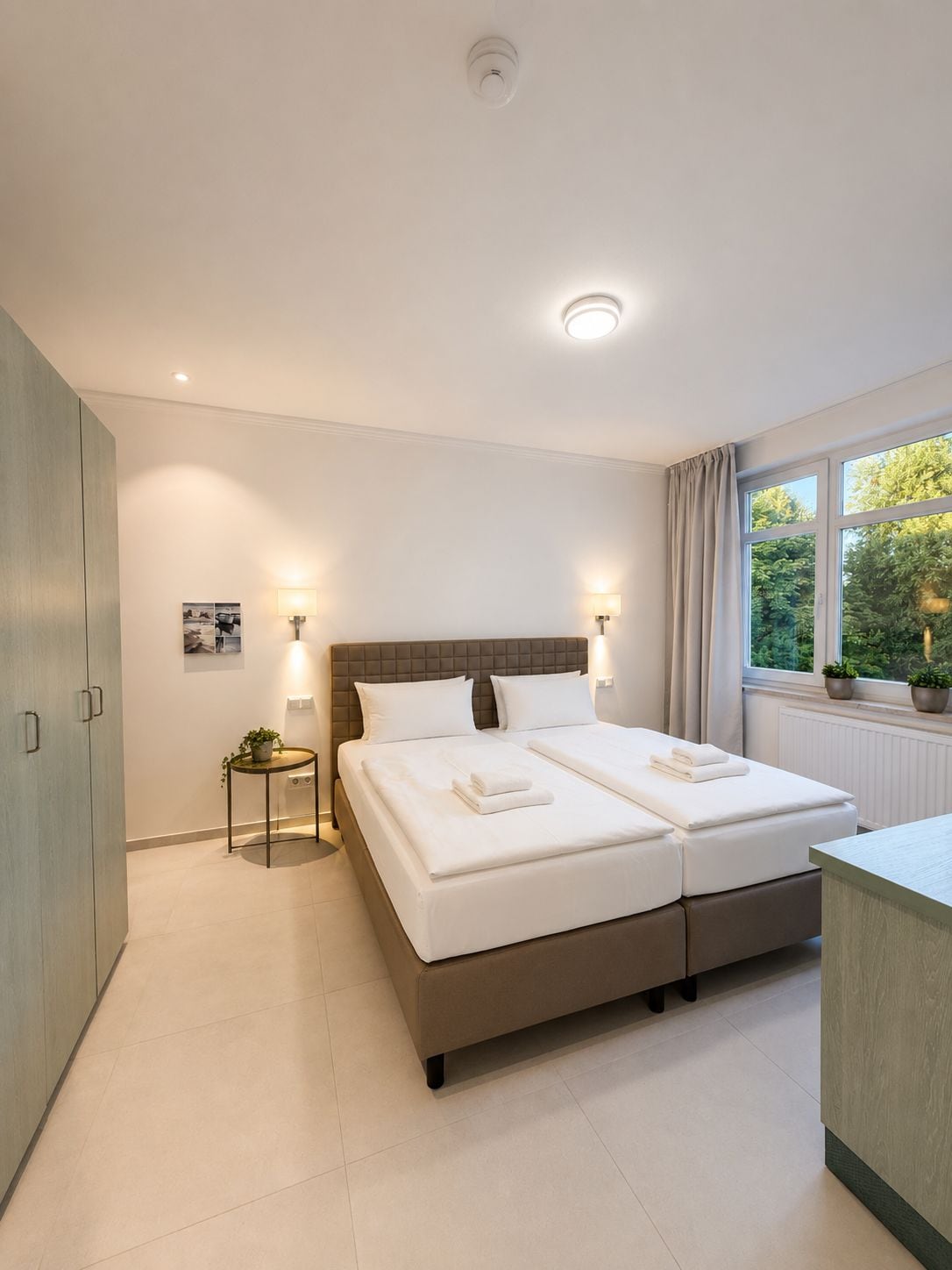 Ein Hotelzimmer mit Doppelbett, Nachttischlampen, einem kleinen Tisch mit Pflanze und einem Fenster mit Blick auf Grünflächen.