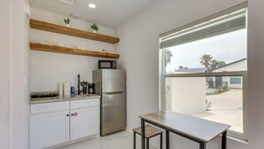 A kitchen with white cabinets, a stainless steel refrigerator, a microwave, wooden shelves, a window with blinds, and a table with chairs. - Beach Base for Groups! SPI Studio w/ Pool On-site (South Padre Island)