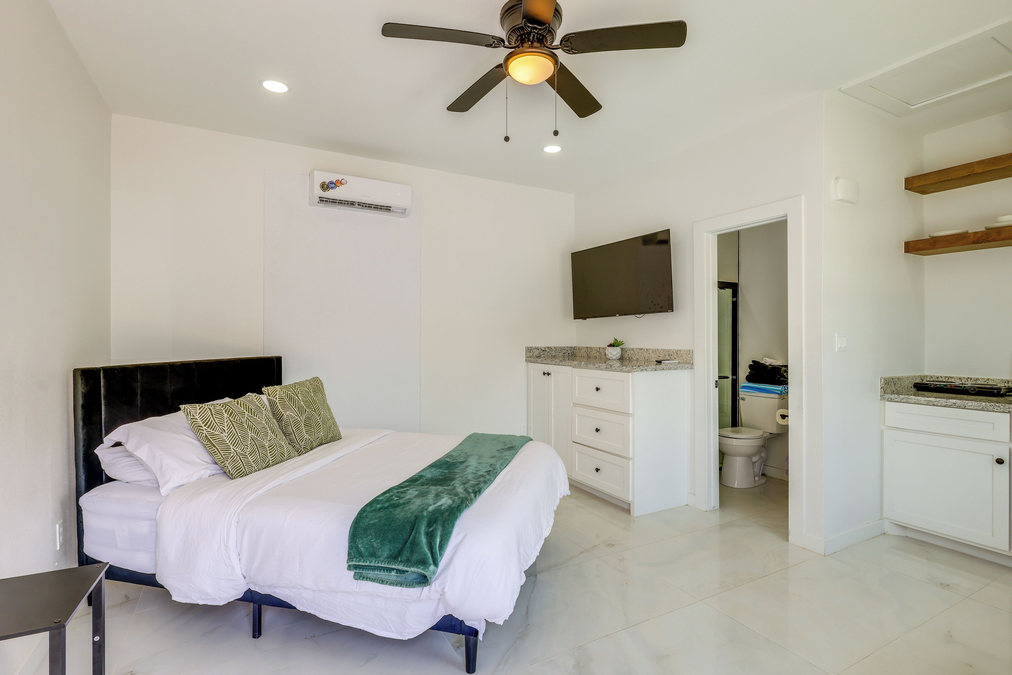 A bedroom with a bed, a ceiling fan, a mounted TV, a white wall air conditioner, and a bathroom visible through an open door.
