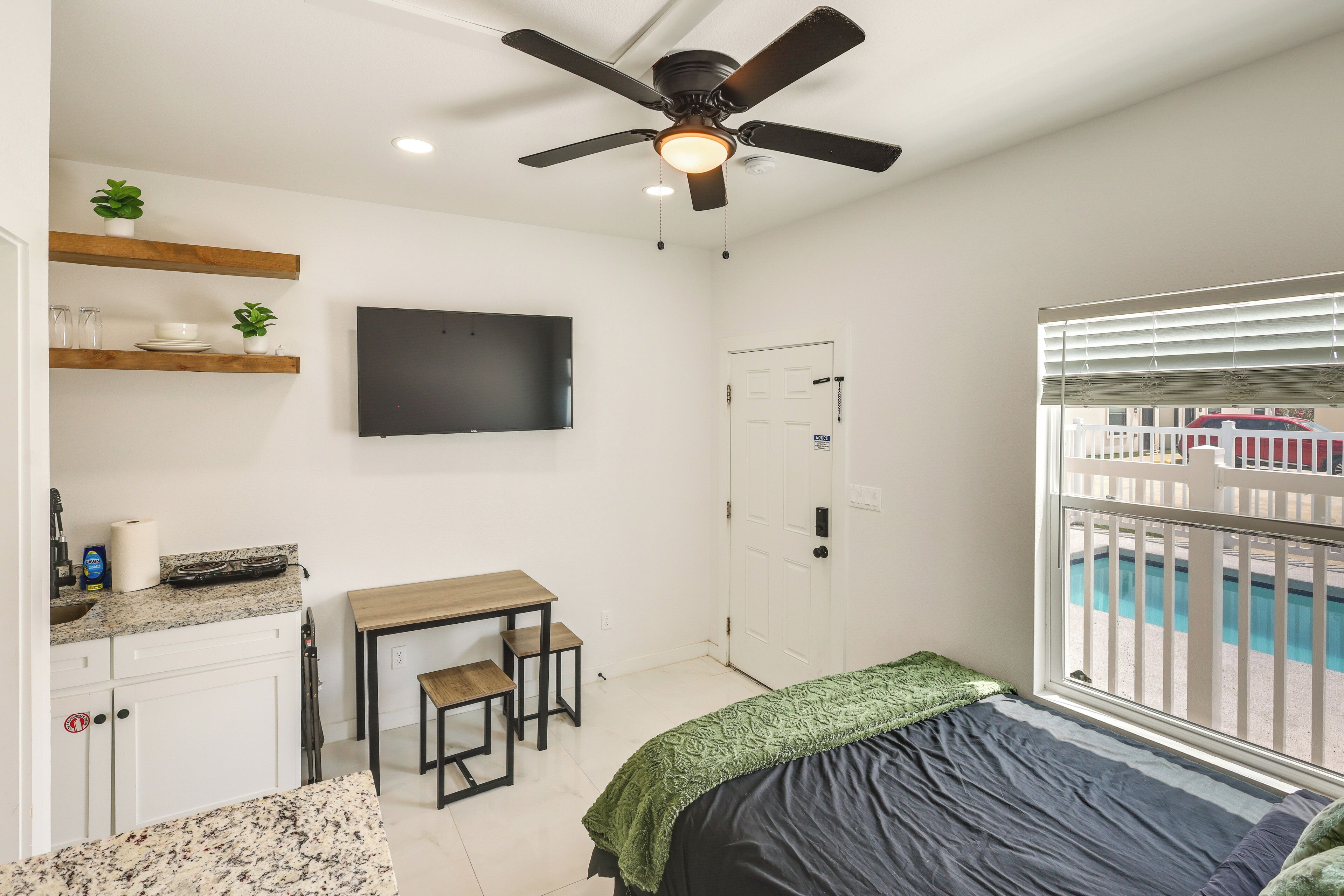 A bedroom with a bed, a desk, a chair, a television, a ceiling fan, and a window with a view of a pool.