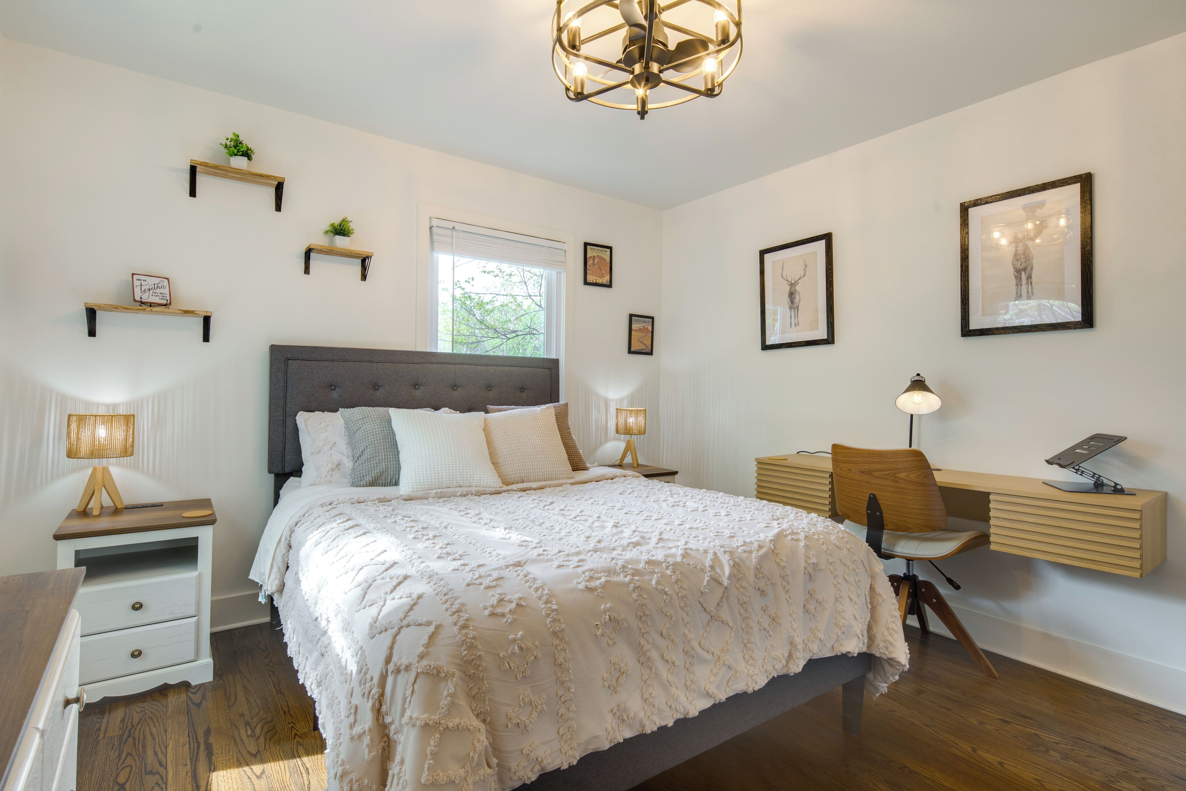 A bedroom with a bed, bedside tables, a desk, and a chair. There are wall-mounted shelves with plants and pictures.
