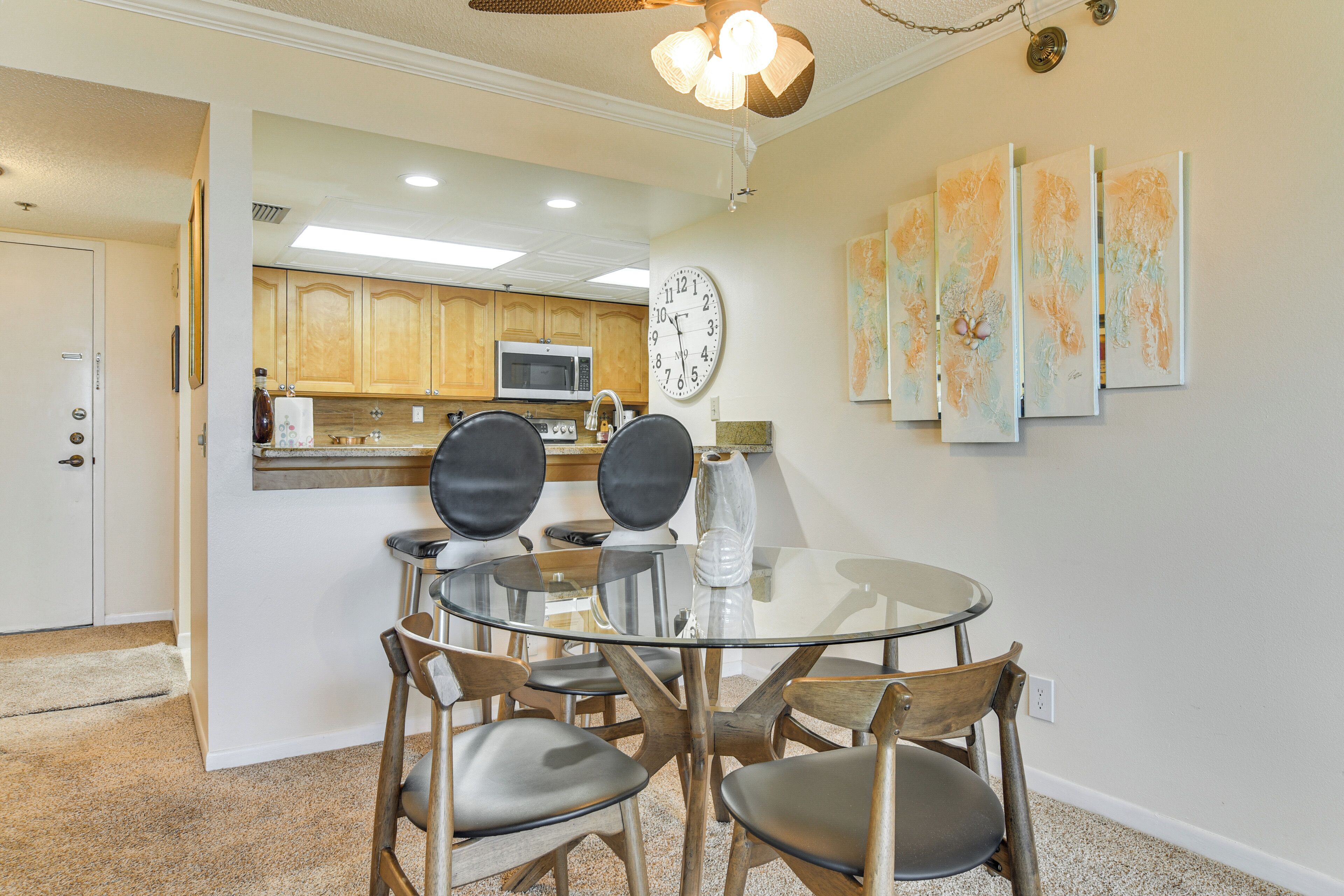 A dining area with a glass table and wooden chairs, a kitchen in the background, and wall art.