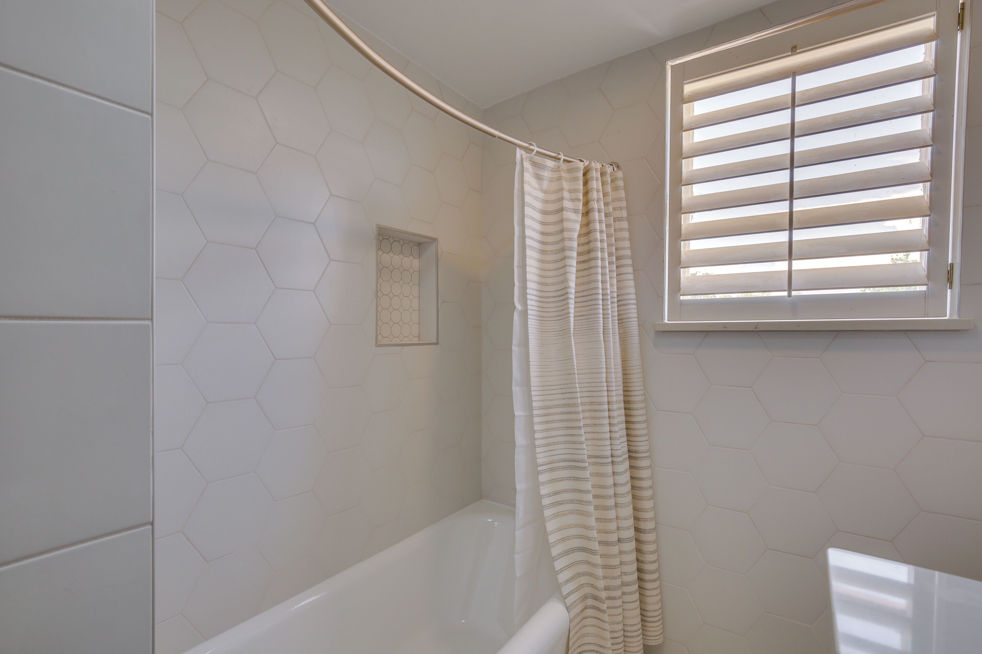 Une salle de bain avec un rideau de douche, une fenêtre avec des stores et des murs recouverts de carreaux hexagonaux.