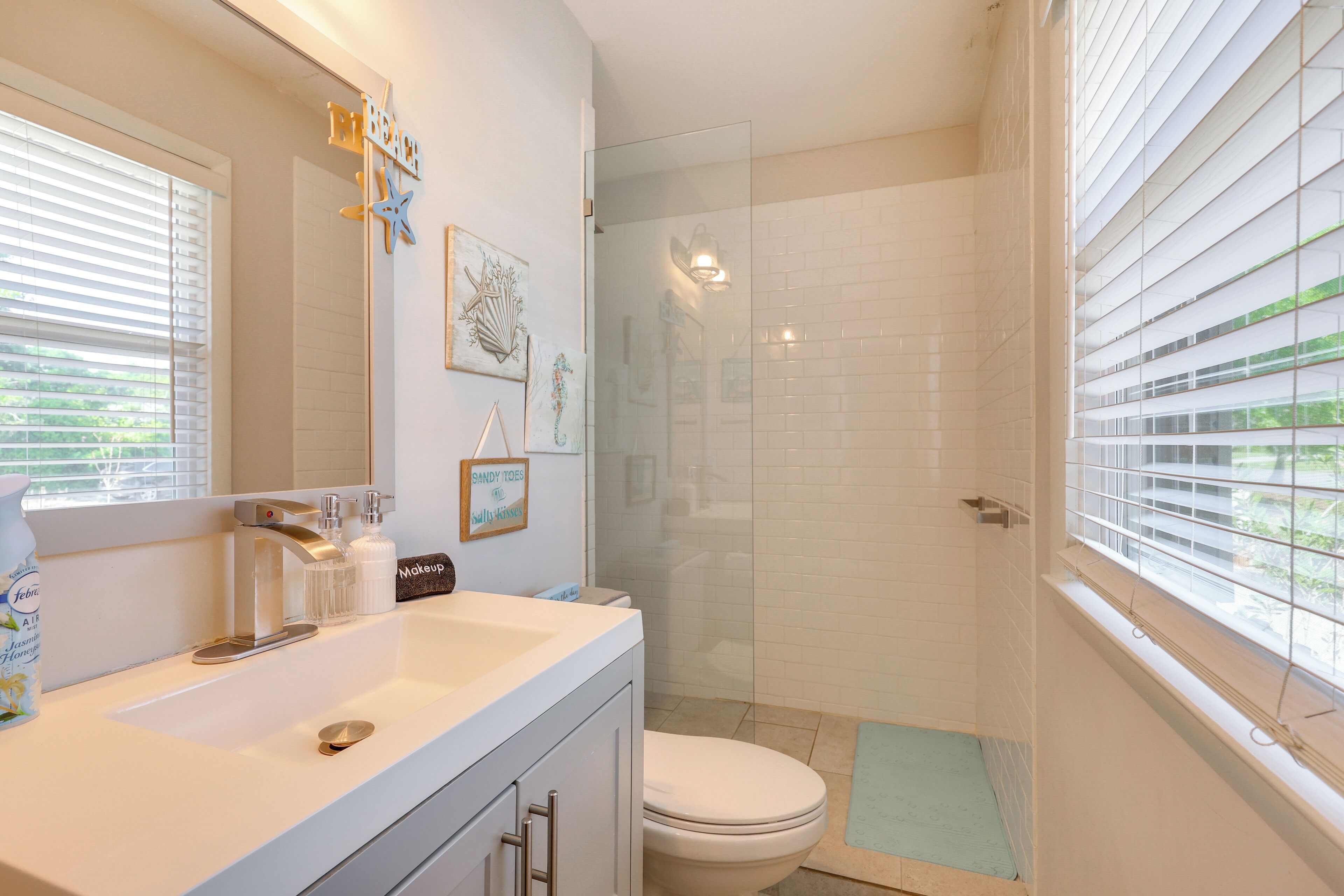 Un baño con lavamanos blanco, inodoro, cabina de ducha de vidrio y ventana con persianas.
