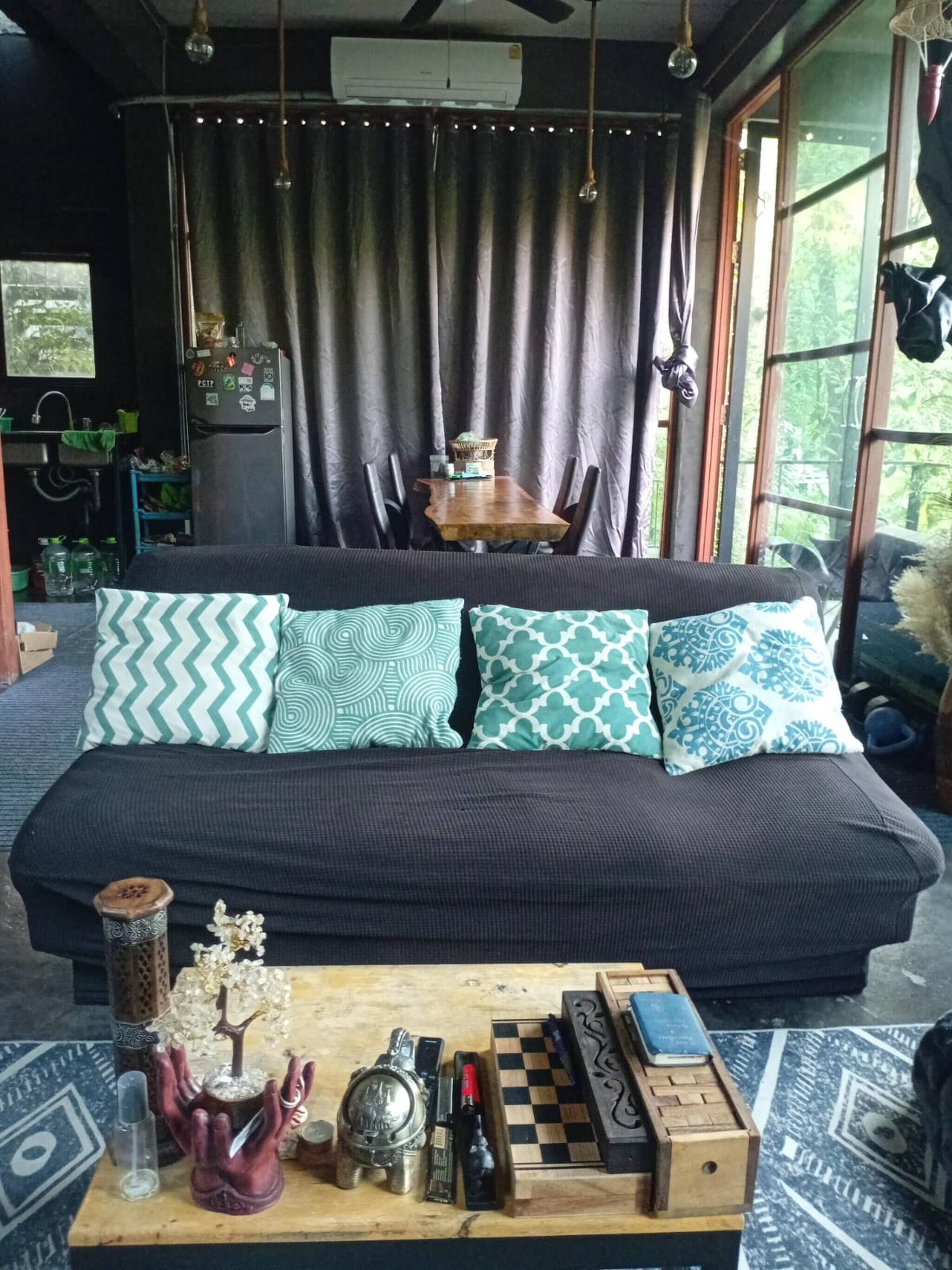 A cozy living room with a dark sofa, patterned cushions, a wooden coffee table with chess, and a view of greenery outside.