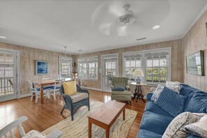 A spacious living room with wooden floors, a blue sofa, a dining table with chairs, and large windows. - The Dolphin House - 4 bedroom soundfront cottage (Wrightsville Beach)