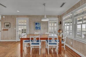 A dining room with a wooden table, white chairs, a hanging light fixture, and large windows. - The Dolphin House - 4 bedroom soundfront cottage (Wrightsville Beach)