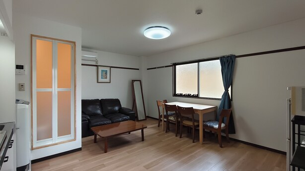 Een woonkamer met een bank, een salontafel, een eettafel met stoelen, een groot raam met gordijnen en een plafondlamp.