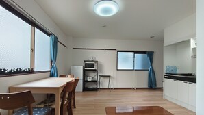 Een kamer met een houten eettafel, stoelen, een magnetron, een kleine koelkast, een raam met gordijnen en een keuken met kasten en een aanrecht.