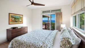 Ein Schlafzimmer mit Bett, Deckenventilator, Nachttisch, Kommode, einem Fenster mit Meerblick und einem Bild an der Wand.