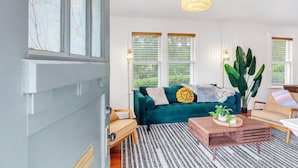 A living room with a teal sofa, a wooden coffee table, and a large plant. - Charming Bungalow Walkable to Main St | King Suite, Yard, Workspace (Elgin)