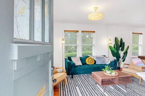A living room with a teal sofa, a wooden coffee table, and a large plant. - Charming Bungalow Walkable to Main St | King Suite, Yard, Workspace (Elgin)