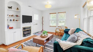 A living room with a sofa, coffee table, TV, and shelves. - Charming Bungalow Walkable to Main St | King Suite, Yard, Workspace (Elgin)
