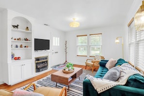 A living room with a sofa, coffee table, TV, and shelves. - Charming Bungalow Walkable to Main St | King Suite, Yard, Workspace (Elgin)
