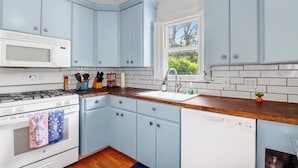 A kitchen with light blue cabinets, a white microwave, a wooden countertop, and a window with a view of greenery. - Charming Bungalow Walkable to Main St | King Suite, Yard, Workspace (Elgin)