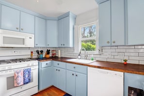 A kitchen with light blue cabinets, a white microwave, a wooden countertop, and a window with a view of greenery. - Charming Bungalow Walkable to Main St | King Suite, Yard, Workspace (Elgin)