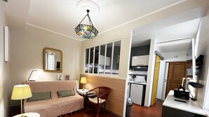 Un espacio habitable compacto con sofá, sillón, una mesita, lámpara, zona de cocina y espejo.