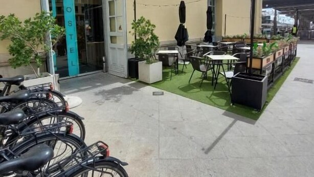 Ostello con un cartello che riporta 'UP! HOSTEL' e una porta verde. Bici parcheggiate all'esterno e una piccola area esterna con tavoli e sedie.