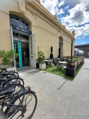 Une auberge de jeunesse avec une enseigne indiquant « UP! HOSTEL » et une porte verte. Des vélos sont garés à l’extérieur, et un petit espace extérieur aménagé avec des tables et des chaises invite à la détente.