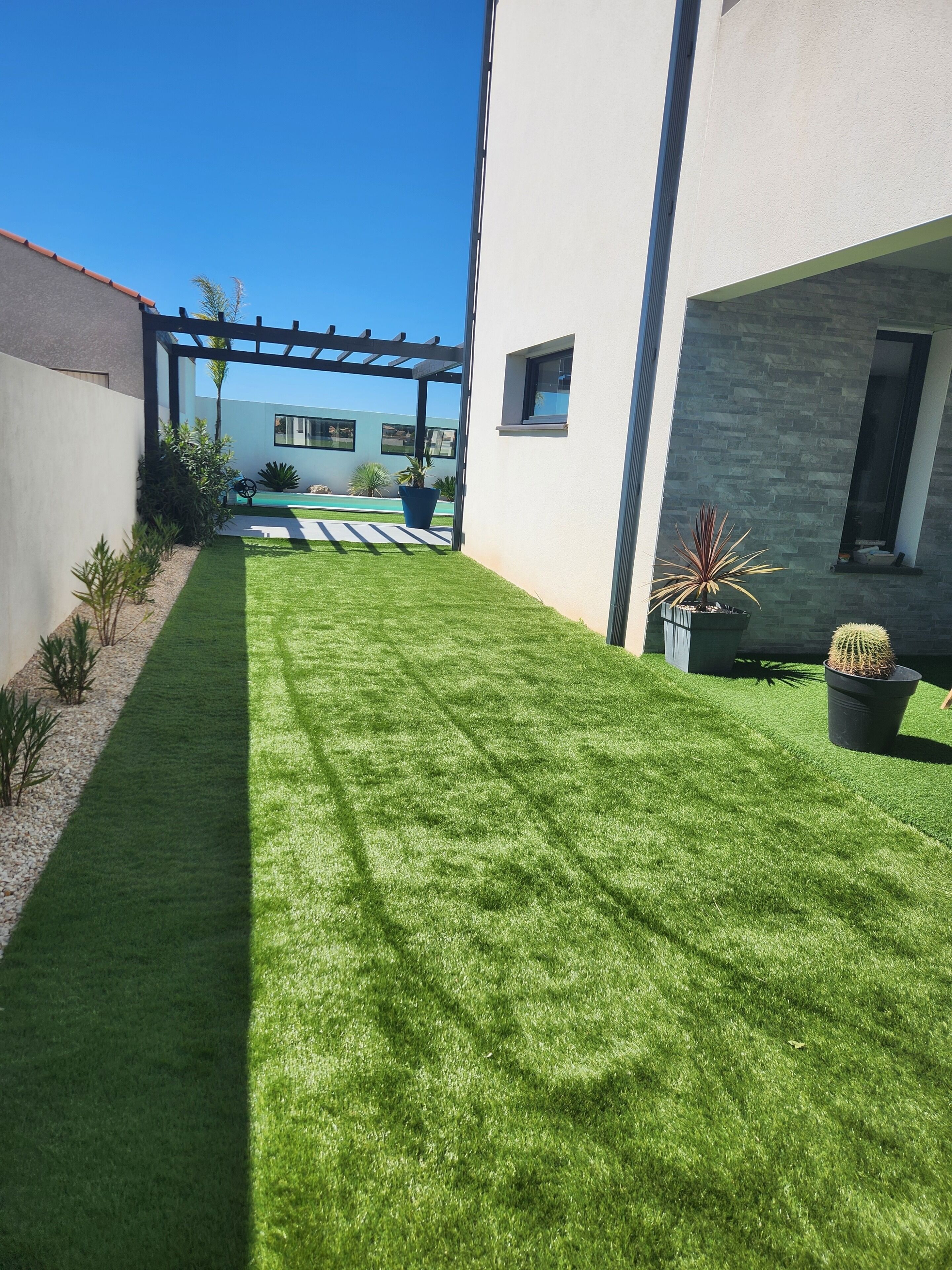Un espace extérieur moderne, aménagé en patio, avec une pelouse bien entretenue, des plantes en pot et une structure en forme de pergola.