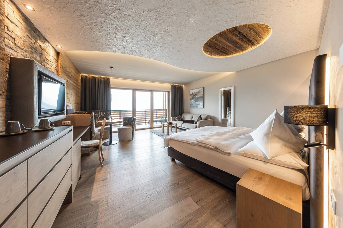 Ein modernes Hotelzimmer mit einem großen Bett, einem Flachbildfernseher, einer Holzkommode, einem Essbereich und einem Balkon mit Blick.