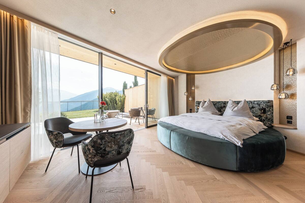Ein modernes Schlafzimmer mit einem großen Bett, einem Esstisch, Stühlen, einer runden Deckenleuchte und Blick ins Freie.