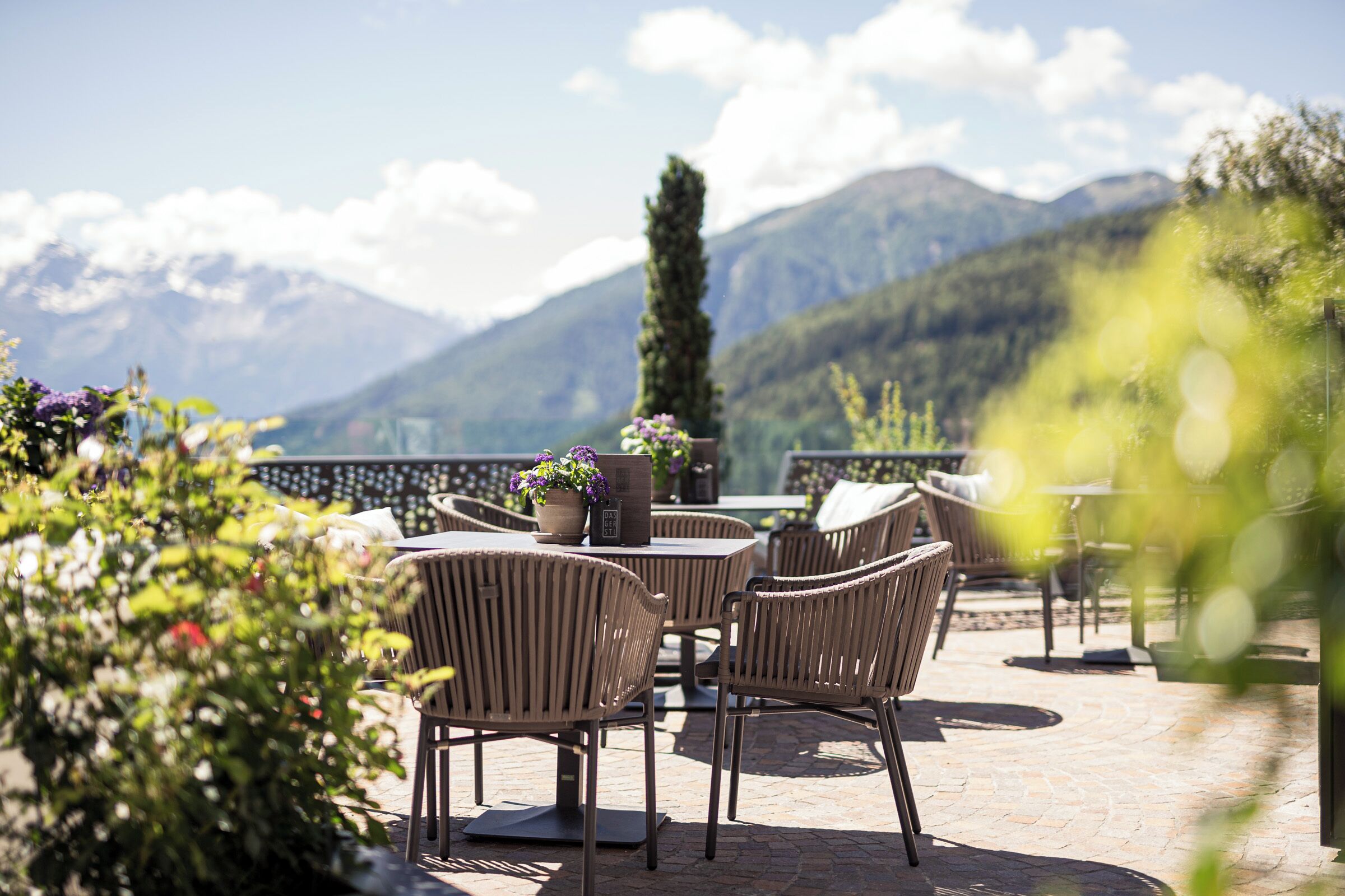 En udendørs terrasse med fletstole og et bord, omgivet af grøn vegetation med udsigt over et bjergrigt landskab.