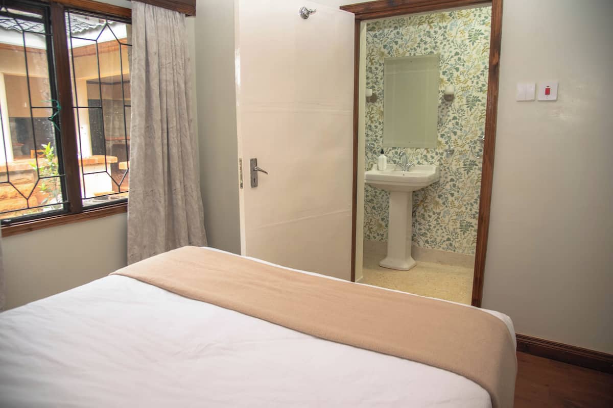 A bedroom with a bed, a window with curtains, a door, and a bathroom with a sink and mirror.